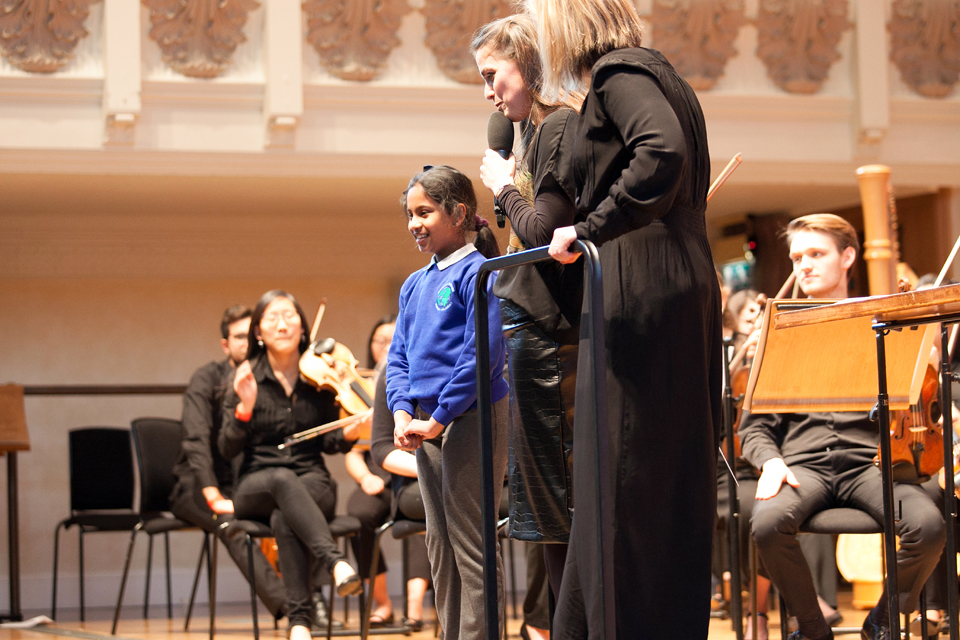 Two women standing, next to a young Asian girl, standing on stage, with an orchestra set up behind them.